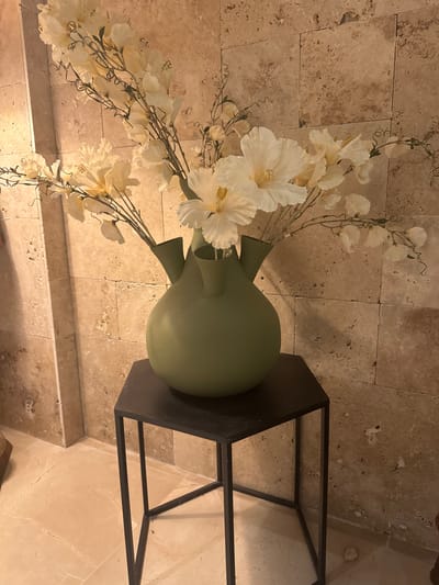 A still life photograph shows a light green ceramic vase with three spouts filled with artificial off-white flowers with delicate stems and leaves. The vase is placed on a small, dark, hexagonal metal table. The background consists of a textured, light brown tiled wall, with one of the tiles appearing to have a patch of white on it. The floor is light-colored with a subtle patterned texture, and there is a faint shadow cast on the wall from the vase and flowers. The image appears to be an interior shot, possibly in a home or establishment in Tirat Karmel, Israel. There are no people or animals visible in the image.
