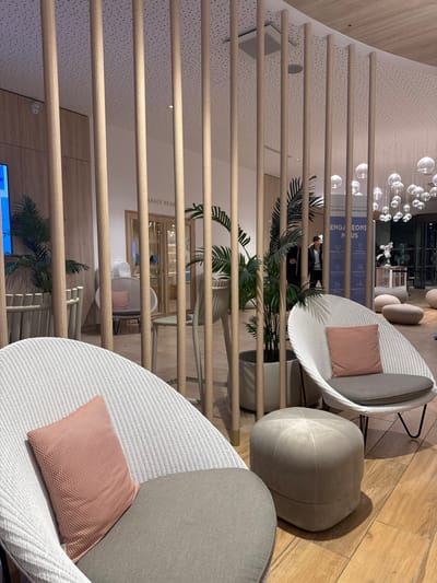 The image displays a contemporary indoor lounge or waiting area, located within a commercial establishment in Marcq-en-Barœul, France.
In the immediate foreground, two white woven armchairs with light grey seat cushions and salmon-pink rectangular throw pillows are positioned. Between these chairs, a light grey upholstered ottoman with a rounded form rests on the wooden floor.
A partition of vertical light wooden slats extends across the midground, creating a semi-transparent divider. Behind this partition, several potted green plants with large leaves are visible. To the left, a portion of a blue digital display screen is present. Towards the center, a light wood-framed doorway or archway is visible, with the partial text "SPACE BEAUTÉ" discernible on the wall above it. To the right of the center, a vertical banner, partially obscured by the slats, displays the French phrase "ENGAGEONS NOUS" in white text on a light blue background.
In the background, a single male individual wearing dark clothing is visible walking towards the right, partially obscured by the wooden slats and a plant. The ceiling features small dark speckles and a suspended light fixture composed of numerous clear glass spherical globes hanging at varying heights. On the far right, several light-colored, organically shaped poufs are arranged on the wooden floor, leading towards a dark entrance or exit point. The floor is covered with light-toned wooden planks. Walls in the background are light beige or white, with some light wood paneling visible on the left.