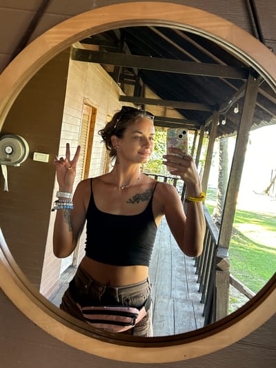 A woman is taking a mirror selfie on a wooden porch in Ban Khlong Hia, Thailand. She is wearing a black crop top, denim shorts, and a pink fanny pack. She has several bracelets on her left wrist and a yellow bracelet on her right wrist. She is holding her phone in her right hand and making a peace sign with her left hand. The porch overlooks a grassy area and palm trees, with the ocean visible in the background. The wooden structure of the porch has exposed beams and a thatched roof. The woman has a tattoo on her chest and arm.