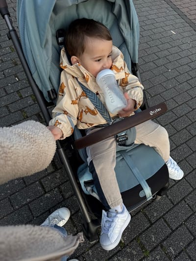 An elevated, outdoor view captures a young child seated in a teal Cybex stroller on a dark grey paver-tiled surface, located in Hanau, Germany. The child, dressed in a beige jacket with brown animal patterns, light-colored pants with dark side stripes, and white Adidas sneakers, is actively drinking from a clear plastic baby bottle containing a white liquid, holding it with both hands. A dark brown padded bumper bar, embossed with the 'Cybex' logo, is positioned across the child's lap. The stroller's multi-point harness system is visible across the child's chest. To the bottom left, a partial view of an adult's body is present, indicated by a cream-colored sherpa-textured sleeve, light blue jeans, and white sneakers, positioned adjacent to the stroller. The environment is illuminated by natural, diffused daylight.