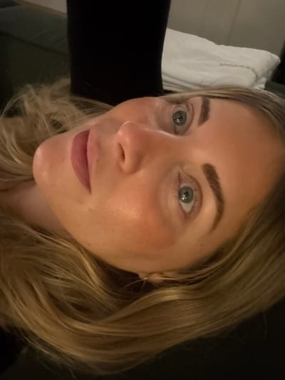 A close-up, high-angle image captures a fair-skinned human subject, appearing to lie down in an indoor setting in Rueil-Malmaison, France. The subject's head is tilted, revealing long, wavy blonde hair framing the right side of the face. Light blue-green eyes, with dark, defined eyebrows, are directed towards the viewer. The nose is central, and the closed lips show a soft pink color, accompanied by a slight blush on the cheeks. The background is blurred, featuring dark fabric to the upper left and a white pillow or folded textile to the upper right. No specific actions or interactions are depicted; the subject is stationary.