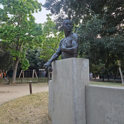 This is a medium shot, eye-level, outdoor photograph of a bronze bust statue in a park. The statue, which depicts a man from the chest up, is positioned on a concrete pedestal. The man has short, dark hair and is looking to his left with his mouth slightly open. His right arm is extended and resting on the pedestal, and he is holding what appears to be a rolled-up scroll or document in his hand. The statue is made of a dark bronze material and has a textured surface.
The concrete pedestal is rough and appears weathered. On the left side of the pedestal, there is some faint text inscribed, but it is illegible.
The park setting is lush with greenery. Behind the statue, there are several large trees with dense foliage. Sunlight filters through the leaves, creating dappled light. To the left of the statue and pedestal, there is a dirt path and a wooden fence. Beyond the fence, there appears to be a playground area with swings and colorful structures. Graffiti can be seen on a wall behind the playground. The ground in the foreground is covered with dry grass and some scattered patches of new green grass. A single, dark brown metal post stands in the grass near the path.
The image appears to be taken in the Parc de la Devesa in Girona, Spain, given the presence of the statue of Enric Marquès and the surrounding park environment which matches descriptions of the park.