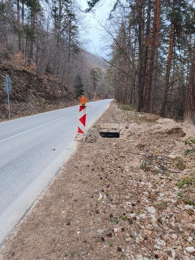 The image depicts a rural road scene in Sveti Konstantin, Bulgaria, characterized by a forested environment and infrastructure damage. The scene is devoid of human presence.
In the foreground, a paved asphalt road extends into the distance, gently curving left. A solid white line marks the right edge of the road. On the immediate right shoulder, a red and white striped warning pole with a flashing amber beacon on top is positioned. A thin, red and white striped caution tape extends from the pole towards the right. Directly behind this pole, an open, rectangular, dark cavity is visible in the ground, consistent with a missing culvert grate or manhole cover. The ground surrounding this opening appears disturbed and consists of loose brown earth, pine needles, dry leaves, and small stones. Further to the right, beyond the open cavity, a mound of disturbed earth and debris is present. A dense line of tall coniferous trees with dark trunks and sparse foliage borders the right side of the road, extending into the background.
On the left side of the road, a sloped hillside rises, covered by a mix of bare deciduous trees and some coniferous trees. A gray road sign with two circular elements is partially visible on a metal pole on the left shoulder. The ground on the slope is covered with dry leaves and sparse vegetation. The sky above the tree line is pale and overcast. No vehicles or individuals are present in the observable scene.