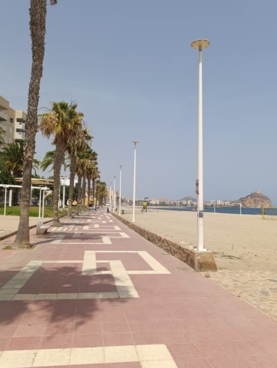 The image depicts a sunny day along a beach promenade in Águilas, Spain. A wide, tiled pathway, decorated with a red and white geometric pattern, stretches into the distance. Palm trees line the left side of the promenade, casting shadows on the path. A low stone wall separates the promenade from the sandy beach to the right. The beach is mostly empty, with a volleyball net visible in the background. Buildings and a small mountain with a fortress-like structure on top form the skyline in the distance. The sky is clear and blue.