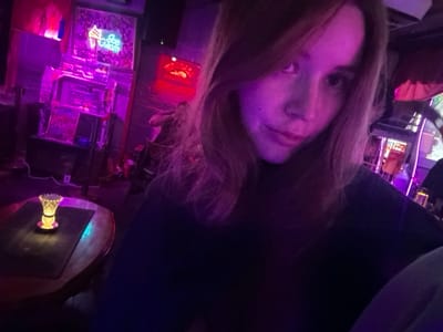 The scene is an interior shot of an establishment located in 마포구, South Korea, characterized by dim, purple and pink neon lighting. A female subject with light brown hair, appearing reddish under the neon light, occupies the foreground, looking directly at the camera with a neutral expression. She is dressed in a dark, high-necked top. To the subject's left, a round wooden table holds a dark placemat and a small, illuminated decorative lamp casting a yellow glow. The background features a wall adorned with multiple neon signs, including a blue 'Ice Cream' sign with an ice cream cone graphic and a partially visible red 'FIREBALL' sign. Below the 'Ice Cream' sign, a multi-tiered clear display case or shelving unit is also illuminated.