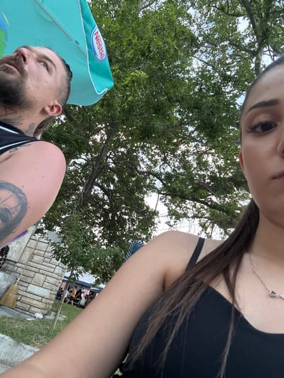 A selfie-style image captures two individuals in an outdoor setting in Kostievo, Bulgaria, beneath a canopy of green trees and a partial view of a blue sky. On the left, a male with a beard, mustache, and visible tattoos on his arm and neck/shoulder looks upwards to his left. He wears a black tank top. A light blue umbrella, with a "Nestlé" logo visible on its underside, partially shades him. On the right, a female, presumably taking the photograph, shows the right side of her face, right shoulder, and upper torso. She has dark hair tied back and wears a black strap top with a silver necklace featuring heart-shaped pendants. In the background, partially obscured by foliage and a stone-clad wall with a broom leaning against it, several indistinct figures are gathered, suggesting a social event or market. The ground shows grass and dirt.