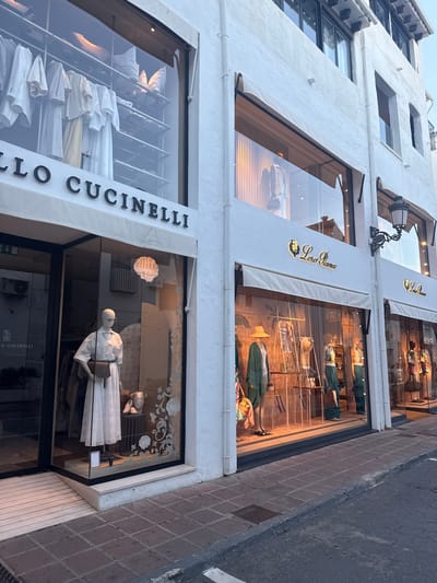 The image shows the exterior of a Loro Piana and Brunello Cucinelli store in Nueva Andalucía, Spain. The facade is predominantly white, with large display windows showcasing clothing and mannequins.
The upper floor display features clothing in light, neutral tones, including what appears to be linen and cotton garments, with mannequins dressed in casual wear such as hats and jackets. The "Loro Piana" logo is prominently displayed on awnings above the windows.
The lower floor display, belonging to Brunello Cucinelli, is also visible. A mannequin is dressed in a white outfit with a textured fabric, holding a handbag. The "BRUNELLO CUCINELLI" name is visible on signage along the side of the window. Inside the store, clothing is neatly displayed on shelves, predominantly in white and light colors.
The scene appears to be set during the day, possibly late afternoon or early evening, given the soft lighting and shadows. The weather seems pleasant, as indicated by the clear sky and the open nature of the store displays. The paving stones outside the store suggest an outdoor, possibly pedestrianized, area. The overall impression is one of a high-end retail establishment in a warm climate.