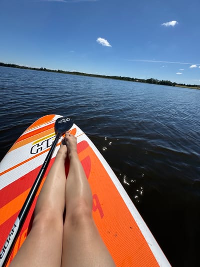 A first-person perspective shot shows a person's legs and feet resting on an orange and white paddleboard in the middle of a dark body of water. A paddle is positioned across the paddleboard, its blade facing upwards. The water is calm with some ripples and reflections from the sun. In the background, a treeline with scattered buildings is visible along the horizon under a bright blue sky with a few white clouds. The image appears to be taken during the day in Lendži, Latvia.