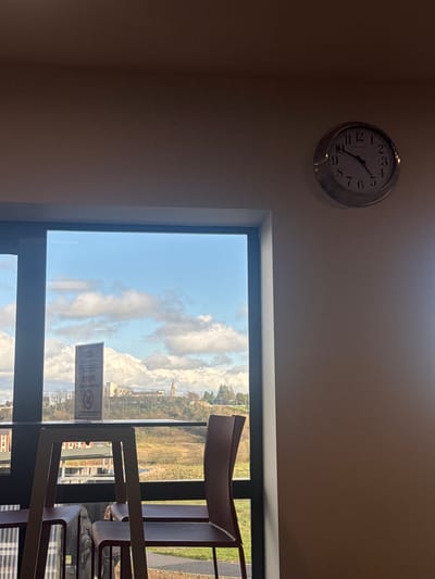 The image presents an interior view through a window, with a wall clock visible above. The clock shows the time as approximately 1:20. The window frames a view of a landscape featuring buildings, green spaces, and a cloudy sky. A sign is affixed to the window, containing text, including the word "STOP!". The focus is drawn to the view outside the window and the elements within the interior space that frame it. Chairs are placed next to the window.
The landscape viewed through the window is in Monaghan, Ireland.