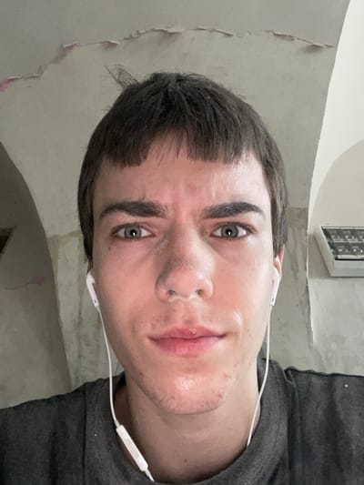 A close-up, frontal, eye-level selfie captures a young person with short, dark hair and pale skin wearing white earbuds and a dark grey t-shirt. The individual has green eyes, prominent eyebrows, and a slightly furrowed brow. The background shows an unfinished interior with exposed plaster on the ceiling, suggesting a renovation or construction site. To the right, a white ventilation unit is mounted on the wall. The lighting is neutral, creating a slightly cool tone. The overall impression is that of a candid, unposed moment. The location is identified as Jihlava, Czechia.