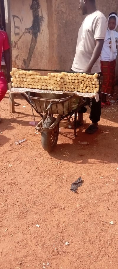 This image captures an outdoor street vending scene in the city of Kasuwan Magani, Nigeria. A metal wheelbarrow, appearing repurposed, supports a vendor's stand draped with a light-colored tarp. On this stand, numerous light yellow, cylindrical pieces of peeled cassava or similar root vegetable are neatly stacked in horizontal rows.
To the right of the stand, an adult male with dark skin stands facing left, wearing a light-colored t-shirt featuring a "Jumpman" logo and dark-colored trousers or track pants, along with black slide sandals. Behind him, a young girl with dark skin is partially visible, wearing a white head covering or hooded top and a patterned long skirt. On the far left, a portion of another individual wearing a red short-sleeved top is visible.
The ground is composed of reddish-brown dirt, with scattered small debris including white paper scraps and a crumpled black plastic bag. In the background, a light brown building wall, showing cracks and wear, features dark graffiti or a painting, including a large letter 'E' and a stylized dark figure. The scene is illuminated by bright natural sunlight, creating distinct shadows.