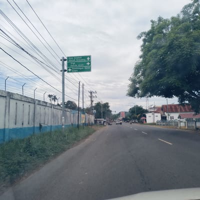 The image captures a street scene in Gampongrayadagang, Indonesia, under an overcast sky. The view is from a vehicle, looking forward along a paved road that curves slightly to the right in the distance. To the left, a long, concrete wall with a blue base and topped with barbed wire runs alongside the road, separating it from what appears to be an industrial or secured area. Overgrown grass and low foliage line the base of this wall.

Prominently featured on the left side of the road is a green road sign mounted on a pole, displaying multiple destinations with directional arrows. The text on the sign reads:
- "Awe Geutah" (Up arrow, indicating straight ahead)
- "Siblah Kreueng" (Up arrow, indicating straight ahead)
- "Sigli" (Right arrow)
- "Banda Aceh" (Right arrow)
- "Bendungan Karet" (Left arrow)
- "Kapa" (Left arrow)

Numerous power lines crisscross the sky above the left side of the road, supported by several utility poles. Further down the road on the left, more structures and smaller buildings are visible, along with additional utility poles and a few parked motorbikes.

On the right side of the road, a large, leafy green tree dominates the foreground, partially obscuring older buildings with corrugated metal roofs further back. The road itself has white dashed lane markings, and several vehicles, including motorcycles and cars, are scattered along its length, suggesting ongoing traffic. The general atmosphere is calm, indicative of a typical daytime street scene in a semi-urban area.