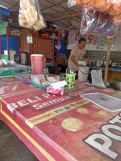 A woman stands at a food stall, holding a white plastic bag. She wears a blue shirt with white lettering and a ponytail. She stands at a counter with a large red plastic container, a small white one, and a stainless steel tea kettle. To the left of the counter, there is a table covered in a bright red tablecloth with the words "Beli Semua One Price" on it. On the table are various snacks and packages of food items. On the table are also plastic utensils in a green holder. The location is in Kecamatan Kartasura, Indonesia, at coordinates (latitude: -7.56, longitude: 110.763). The environment is a food stall with a wooden frame, and the scene is lit by natural sunlight. There is an open sky above the stall. The entire scene is contained in a single image.