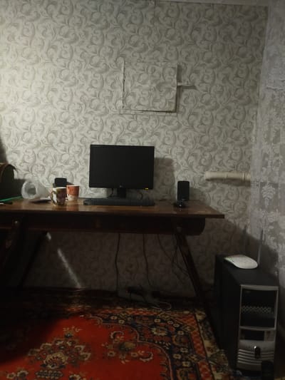 The image depicts an indoor scene, likely a home office or study, with a computer setup. A wooden desk is centrally placed, supporting a computer monitor, keyboard, and mouse. On the desk, there are two mugs and what appears to be a plastic bag. A computer tower is visible to the right of the desk, topped with a wireless router. The walls are covered in patterned wallpaper, and a traditional rug with a red base and intricate designs lies on the floor. A pipe runs horizontally along the wall behind the desk, and a white curtain hangs on the far right. The lighting suggests it is nighttime or a dimly lit room, with a beam of light cast on the rug. The image does not provide any specific indicators of the city of Martuk, Kazakhstan, or any other geographical location. There are no people or animals present in the scene, and no discernible actions or interactions are taking place.