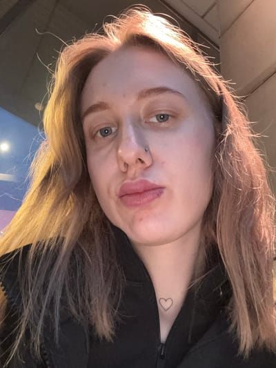 A selfie of a fair-skinned person with light brown hair. The person has blue eyes, a piercing on the left side of their nose, and a small heart tattoo on the neck. The person is wearing a black jacket. The location appears to be indoors, with a blurry blue and pink surface visible in the lower part of the frame. No other people or objects are identifiable in the image. There is no indication this image was taken in Ol'ginskaya, Russia.