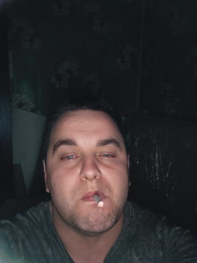 A close-up, self-portrait photograph shows a fair-skinned male with dark hair and stubble, holding a lit cigarette in his mouth. He is wearing a dark gray t-shirt. The background is dark and appears to be a wall with a floral pattern. The lighting is dim and appears to be artificial. The image appears to be taken indoors. There is no visible indication of the city of Ketrysanivka, Ukraine.