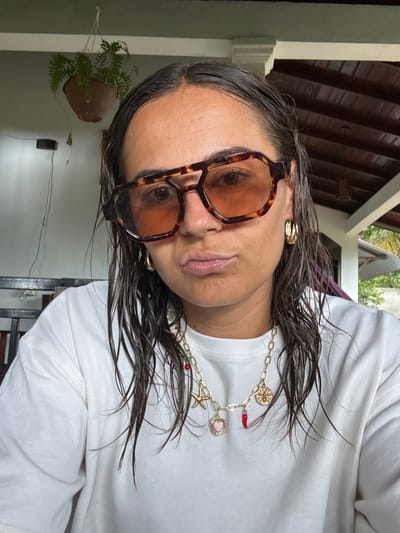 A female subject is depicted in a close-up, selfie-style portrait within a semi-outdoor setting in Nilwella, Sri Lanka. She has dark, damp hair parted centrally and an olive complexion. Her lips are pursed in a pout. She wears large, tortoise-shell, hexagonal-aviator style sunglasses with amber-tinted lenses. A gold-toned hoop earring is visible on her right ear. Around her neck is a gold-toned chunky link necklace adorned with multiple charms, including a gold starfish, a pink heart with a gold border, a red chili pepper, and a gold medallion. Small red and green beads are also integrated into the necklace chain. She is wearing a plain white crew-neck shirt. The background on the left features a light blue painted wall with a hanging terracotta pot containing a green fern plant. A dark wooden or metal furniture piece is partially visible below the plant. The background on the right shows a wooden slatted ceiling with visible rafters and a white painted beam, alongside distant green foliage and a glimpse of sky, indicative of an open veranda or patio environment.