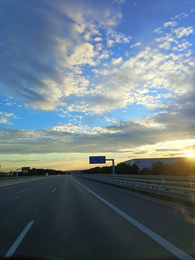 This is an image of a highway in Germany, likely near Ulm, with a sunset or sunrise in the background. The perspective is from the driver's seat of a vehicle, looking forward. The road is asphalt with white lane markings. On the right side of the highway, there is a metal guardrail and trees. In the distance, there is a large building with "SDLL" visible on it, possibly a warehouse. On the left side of the highway, there are more trees and some smaller signs. The sky is filled with clouds, with the sun glinting brightly on the right side of the frame. Traffic appears light, with only a few vehicles visible in the distance. A blue highway sign indicating directions to Stuttgart, Ulm, and Geislingen is visible on the right.
