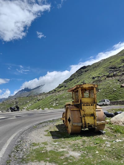 The image depicts a scenic mountain road in Yari-Khoksar, India. In the foreground, a weathered yellow road roller is parked on the grassy verge next to a paved road. The road curves to the left, with a black scooter parked on the opposite side. Beyond the road, a steep, green, rocky mountainside rises towards a bright blue sky dotted with white clouds. In the distance, more majestic mountains are visible, partially shrouded in mist. A white SUV is parked further up the road, near a stone wall. The environment appears to be a high-altitude, mountainous region, likely on a clear day with some cloud cover.