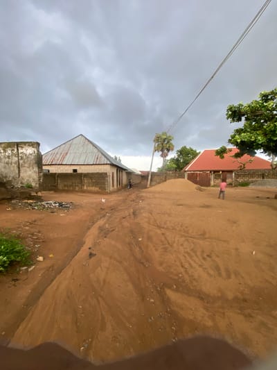 A dirt road in Jere South, Nigeria, is the scene in this image. The sky is overcast with gray clouds. On the left, a partially constructed building with a corrugated metal roof and concrete block walls stands next to a dilapidated wall covered in graffiti. Trash is piled near the building. Further down the road, another building with a red tile roof and wooden doors is visible. A young boy in a pink shirt stands near this building, facing away from the camera. A palm tree is also present, and power lines stretch across the sky. The road is covered in red dirt, with some evidence of erosion and water flow.