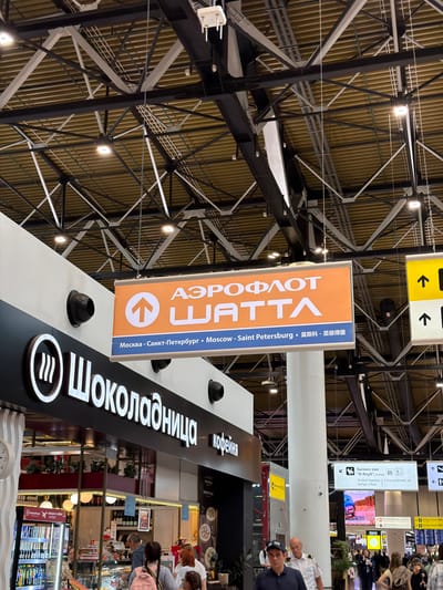 The image appears to be taken inside an airport terminal in Perepechino, Russia. The focal point is a prominent orange and blue sign with Russian text and an upward-pointing arrow, indicating Aeroflot Shatra airline. Below this, text reads "Москва - Санкт-Петербург - Moscow - Saint Petersburg - 莫斯科 - 圣彼得堡", suggesting destinations served by the airline. To the left, a sign for "Шоколадница" (Shokoladnitsa), a Russian coffee shop chain, is visible, with people queuing and browsing items in the display. Further into the background, there are directional signs, including one for "Бизнес-зал" (Business Lounge) and another with arrow icons. The ceiling structure is a complex network of metal beams and yellow corrugated panels, typical of airport architecture. Several people are walking through the terminal, some with luggage, indicating travel activity. The overall environment is brightly lit with artificial lighting.