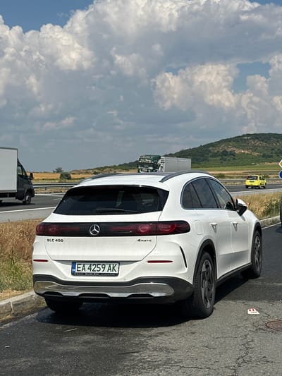 The image shows the rear of a white Mercedes-Benz GLC 400 4Matic parked on the side of a highway. The car has a Bulgarian license plate reading "EA 4259 AK". In the background, there is a truck and a yellow car on the highway, with a hilly landscape and fields on either side. The sky is partly cloudy. The scene is located on a highway in or near the city of Drazhevo, Bulgaria, with dry vegetation visible along the roadside. The tarmac on the road is worn and cracked.
