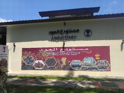 This image depicts the exterior of a building located in Nová Bystřice, Czechia. The building features a cream-colored facade with a dark brown roof overhang. Affixed to the wall above a large maroon banner are two signs: one displaying Arabic script "مستودع العود" and English text "Abdul Hady," and an adjacent circular sign containing a stylized, abstract emblem within a dark border. Two black wall-mounted lanterns are visible on the facade, one on the left and one on the right.
The prominent maroon banner is richly decorated with intricate gold and red patterns, a crescent moon, and two stylized lanterns. It features Arabic script "مستودع العود" at the top, followed by English text "Abdul Hadi Oud" and "PT. MEDINA ABDUL HADY GROUP." Below this text, six hexagonal frames showcase various images: two piles of dark, raw materials (possibly wood chips or resins), a cityscape, another pile of dark materials, a display of bottles containing colored liquids (green, pink, yellow), and a collection of what appear to be refined products or ingredients.
The foreground consists of a paved path with sections of lighter and darker gray, bordered by green grass. A portion of a tree trunk is visible on the far left. The scene is illuminated by natural light under a clear blue sky, indicating fair weather conditions. No individuals or active interactions are visible.
