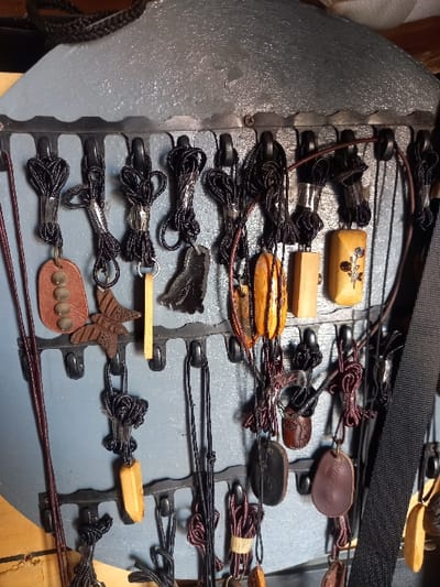 The image shows a display of various handcrafted items hanging on a metal rack, likely for sale. The rack is mounted on a wall painted a light shade of blue, with a dark brown wooden frame surrounding the display. The rack is made of metal bars with hooks along its length.
Various items, including pendants, necklaces, and charms, are hanging from the hooks. The pendants, primarily crafted from wood and featuring carvings, are in shades of brown and dark brown. Some have intricate designs, while others are plain. The majority of the hanging items are attached to black cords, but some are secured with brown or black leather straps.
The image suggests a local market or craft shop in Ita Oniyan, Nigeria. The items likely represent traditional craftsmanship and are potentially sold to locals or tourists. The precise location cannot be determined from the image alone, but it depicts an interior space with the potential for a storefront.