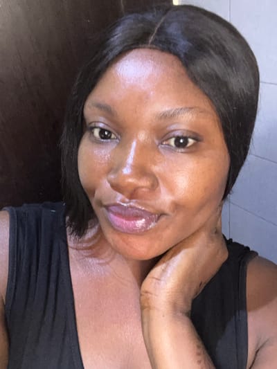A close-up, chest-up portrait shows a female individual in Dzhidu, Nigeria, against an indistinct indoor background. The subject has dark skin, dark, straight hair parted in the center and reaching approximately shoulder length. She is wearing a dark, sleeveless garment. Her face exhibits a glossy appearance, particularly on the forehead, nose, and cheekbones. Her lips are full and coated with a light pink or purplish gloss. Her dark eyes are open and directed towards the viewer. The individual's right hand is visible, with fingers resting against her neck and lower jawline. The background consists of a dark, uniform surface on the left and a lighter, potentially tiled or painted wall on the right.