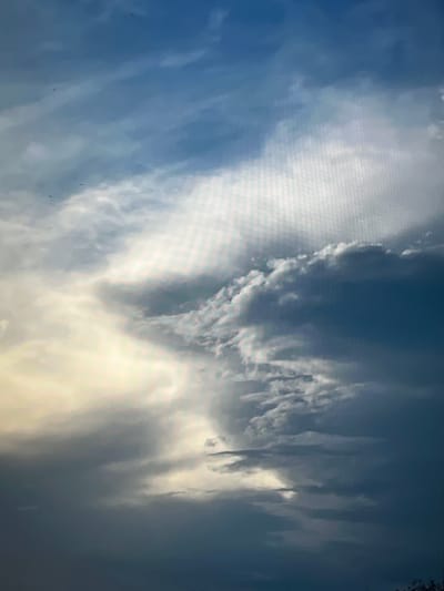 This is a close-up, low-angle photograph of a cloudy sky, taken on a day that is likely either before or after a storm. The sky is a mix of dark grey, deep blue, and bright white clouds. Sunlight is breaking through some of the clouds, creating a dramatic, almost ethereal effect, with beams of light illuminating portions of the sky. The overall mood is atmospheric and somewhat somber, but also beautiful. There are no discernible features of the city of Yenakiieve, Ukraine, in this image, as it is solely focused on the sky. The image does not contain any people, subjects, or objects other than clouds, nor does it depict any actions or interactions.