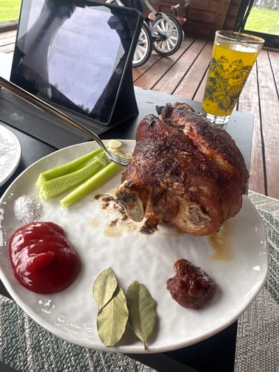 A close-up, eye-level shot shows a plate of roasted pork knuckle with accompaniments on a wooden deck. The pork knuckle is prominently displayed, with crispy skin and visible bone structures. To the left of the pork are several sticks of cucumber, a small pile of white salt, and a dollop of red ketchup. Two dried bay leaves and a small mound of a dark reddish-brown sauce, possibly mustard, are situated on the lower right side of the plate. In the background, a glass of yellow liquid with a decorative pattern is visible, along with a tablet device propped up on a stand. The setting appears to be outdoors on a weathered wooden deck, with the blurry background suggesting a natural or semi-urban environment. The image is taken in Борисьево Днп, Russia.