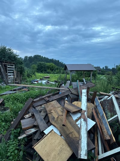 In a rural outdoor setting, likely Gon'ba, Russia, a large pile of discarded lumber and building materials dominates the foreground. The materials are assorted in shape, size, and condition, with some pieces appearing weathered and others newer. The lumber is densely packed and jumbled, creating an uneven terrain within the frame.
Behind the pile of debris, the environment transitions to a natural landscape. Lush green vegetation, including tall grass and bushes, surrounds the lumber. In the mid-ground, a simple, rustic wooden structure with a metal roof stands to the right, possibly a shelter or wellhead. Further in the background, a dense line of trees forms a treeline against a cloudy, overcast sky. The overall impression is one of neglect or a site undergoing demolition or construction cleanup in a rural area. No people or animals are visible in the image. The lighting suggests it is late afternoon or early evening, or the day is heavily overcast.