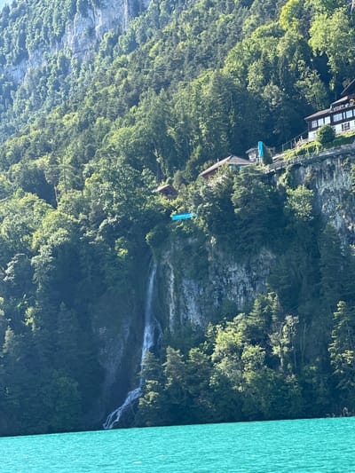 In Sundlauenen, Switzerland, a turquoise lake is in the foreground. Behind the lake, a steep, forested cliff rises. A waterfall cascades down the cliff face. Several buildings with brown roofs are nestled amongst the trees on the upper part of the cliff. A blue banner is visible on the cliff face near the top of the waterfall.