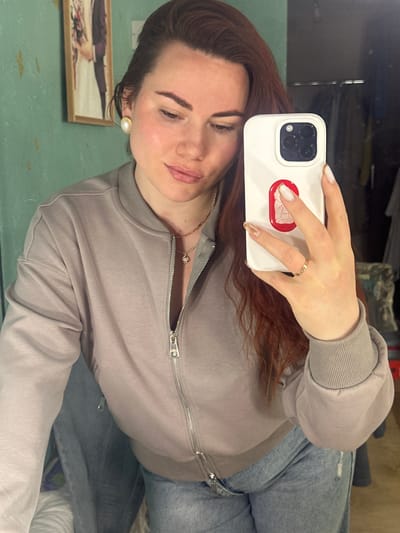 A woman is taking a selfie in what appears to be a room or apartment, possibly in Черёмушки, Russia, although this cannot be definitively confirmed without additional information or context.
The woman has reddish-brown hair and is wearing a light gray zippered jacket, a delicate necklace, and a pearl earring. She is holding a white smartphone with a distinctive red and white design on the case, and a gold ring is visible on her finger. She has light skin and is wearing makeup.
She is wearing blue jeans with what appear to be distress marks. Behind her and the woman in the frame is a painting on a light green surface. Parts of furniture/bedding are visible in the background. A reflection is seen in the top left corner.