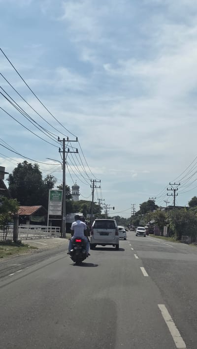 The image depicts a street scene in Ketapang, Indonesia, taken from the perspective of someone traveling along the road. In the foreground, a motorcycle with a rider wearing a white shirt is moving in the same direction as the camera. In front of the motorcycle, a white van travels ahead, followed by other vehicles in the distance. Overhead power lines crisscross the sky. On the side of the road, there is a sign that reads "Apotek Sabina". A building with a white dome is visible behind some trees and the Apotek sign. The sky is light blue with scattered clouds. The road is asphalt with a broken white lane marker.