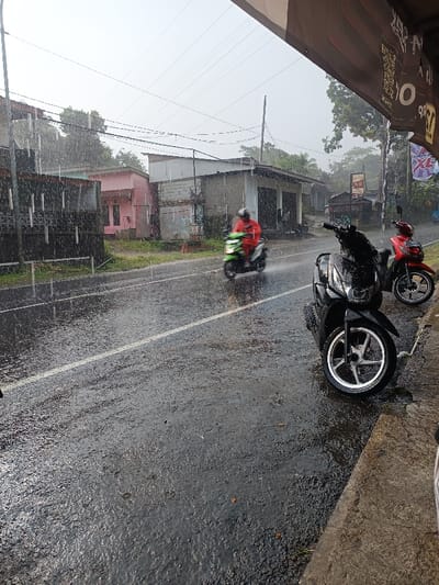 The image captures a rainy daytime street scene in Cugenang, Indonesia. Heavy rainfall is actively occurring, visible as streaks across the frame and saturating the dark asphalt road, which reflects the overcast sky. In the foreground on the right, a dark grey concrete curb runs horizontally. Two motor scooters are parked parallel to this curb, facing away from the viewer: the closest is a black scooter with silver-colored five-spoke wheels, and behind it is a red scooter. Above these, a brown awning, partially visible in the upper right, displays black and white graphics. In the midground, a person wearing a red raincoat and a dark helmet is riding a light green motor scooter away from the camera on the wet road, with the rear wheel generating a visible spray of water. A continuous white line marks a lane on the asphalt. The background on the left features a series of residential or commercial buildings, including a light pink structure and a grey concrete block building, interspersed with green trees and dense foliage. Utility poles and numerous overhead power lines are visible against the diffused light of the sky.