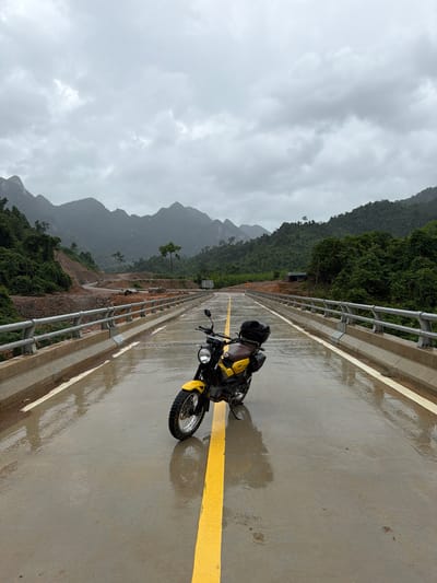 A yellow motorcycle is parked on a wet, empty bridge in Trường Sơn Commune, Vietnam. The road is slick with water, reflecting the overcast sky and surrounding mountains. A bright yellow line divides the road, and the motorcycle is positioned directly on it. The bridge has metal railings on either side, and beyond them, steep, densely forested mountains rise into the cloudy sky. Construction or road work is visible on the left side of the road leading up to the bridge. The scene evokes a sense of solitude and the rugged beauty of the Vietnamese landscape, possibly during or after rainfall.
