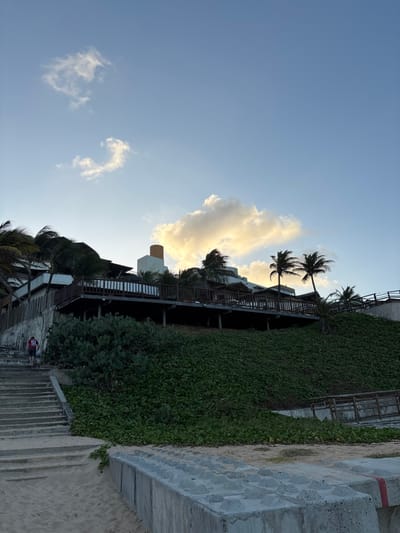 The image depicts a coastal scene in Natal, Brazil, at either sunrise or sunset, characterized by a pale blue sky with scattered white and gold-tinted clouds. A prominent multi-story building, likely a resort or residence, is situated on a steep, vegetated incline in the midground. The building features white walls, a distinctive light-brown cylindrical chimney-like structure on its roof, and multiple wooden balconies or decks with dark railings. Numerous palm trees, some appearing wind-swept, are interspersed around the building and along the slope.
The incline itself is densely covered with dark green ground vegetation and some larger, possibly cacti, shrubs near its base. A weathered concrete staircase ascends the left side of the slope from the sandy foreground towards the building. One individual is visible on these stairs, positioned approximately halfway up, walking away from the viewer. This person is wearing a red or orange t-shirt with a white number "51" on the back and dark shorts.
The foreground consists of light-colored sand, partially obscured on the right by a concrete retaining wall or barrier. This barrier has a textured top surface with regularly spaced protrusions and a faint red stripe visible on its side. No direct interactions between individuals are depicted, and the overall scene conveys a calm, outdoor environment.