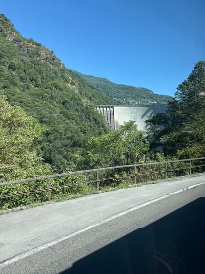 The image shows a large concrete dam in a mountainous, forested area. The dam is curved and has a series of vertical structural elements visible. The surrounding landscape is covered in lush green trees, with steep rocky slopes on the left side. The sky is clear and blue. In the foreground, there is a paved road with a white line marking its edge, and a metal railing separates the road from the vegetated area below. A shadow of an object, possibly a vehicle, is cast across the road. The location is identified as the city of Gordemo, Switzerland.