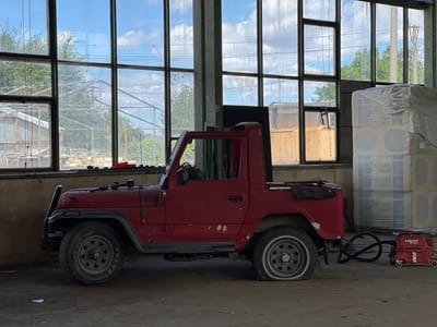In Lanžhot, Czechia, a red, open-top Jeep-style vehicle with a flattened rear tire is parked inside a large industrial building. The building has a concrete floor and large, grimy windows that let in daylight, revealing trees and a cloudy blue sky outside. To the right of the vehicle, a red Hilti VC 20X vacuum cleaner is connected to the vehicle by a black hose. Stacked white pallets, possibly of insulation material, are visible behind the vacuum cleaner. The Jeep appears to be in a state of disrepair, with visible rust and minor damage to its exterior. The overall scene suggests a storage or workshop area within an industrial setting.