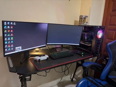 The image displays an indoor computer workstation situated in Icod de los Vinos, Spain. A black L-shaped or corner desk is the central feature, supporting a dual-monitor setup. The left monitor displays a desktop environment with numerous application icons and a red crosshair, while the right monitor presents a dark screen. A white PlayStation 5 DualSense controller rests on a black mouse pad with red stitching, positioned directly in front of a black gaming keyboard and a black Razer gaming mouse.

To the right of the monitors, a black PC tower with a visible interior showcases components such as a CPU cooler and a "GeForce RTX" graphics card, illuminated by multi-colored RGB fans and lighting. A small, warm-toned lamp is visible beneath the left monitor. The background consists of a light-colored, textured wall. In the upper right corner, a yellow-themed Funko Pop figure is placed on a shelf. A brown wooden door is partially visible along the far right edge of the frame. A blue and black gaming chair is positioned at the desk, with its armrest prominent in the lower right foreground. No individuals are present in the scene.