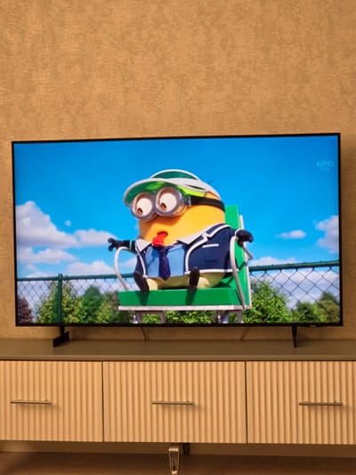 A television screen displays an animated scene featuring a minion character wearing a blue suit, white shirt, and tie, and a green visor with goggles. The minion is seated on a green chair with a white frame, its tongue sticking out. The background shows a clear blue sky with white clouds, a chain-link fence, and green trees. In the upper right corner of the screen, the text "КИНО ГИД" is visible. The television is placed on a stand in front of a textured, light brown wall. Below the television is a light-colored cabinet with fluted drawer fronts and silver handles. The image captures an indoor scene, likely a living room, where the television is turned on. The presence of "КИНО ГИД" suggests the television is likely in Russia, possibly in the city of Komsomolskoye, Russia.