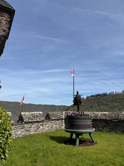 The image depicts an outdoor scene in Cochem, Germany. A large, wooden wine press, repurposed as a flower planter, stands on a well-maintained green lawn beside a stone wall. Flowers in white and purple tones adorn the top of the press. A metal rod extends upwards from the press, capped with a small, red and white flag. In the background, a wooded hillside stretches towards the blue sky with wispy clouds. Another red and white flag is visible in the distance on the upper left of the screen, flying from the wall. The stone wall, constructed of unevenly sized stones, varies in height and borders the lawn. A fragment of stone architecture is visible at the very bottom left of the screen.