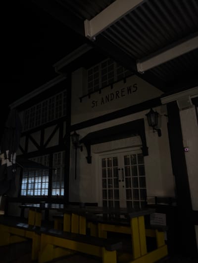 This nighttime image depicts the exterior of "ST ANDREWS" in Port Alfred, South Africa. The building exhibits a Tudor-style facade with white textured walls and dark horizontal and diagonal timber-like beams. A dark, corrugated metal roof extends over the structure. The name "ST ANDREWS" is displayed in dark capital letters on a light background above a central white double door entrance, which features multiple glass panes.

Two black, wall-mounted lanterns illuminate the facade, positioned to the left and directly above the main doors. To the right of the entrance, a small security device or camera with a purple indicator light is affixed to the wall.

In the foreground, an outdoor seating area is visible, consisting of multiple picnic-style tables with integrated benches. These tables feature bright yellow supports and dark-colored tops. To the far left, a large, dark patio umbrella is seen in a collapsed and tied state. A small white sign with dark text is partially visible near the tables on the right, displaying "PLEASE MAKE USE OF THE BIN" and "TO DISPOSE OF" with further text obscured. No individuals are present in the scene.