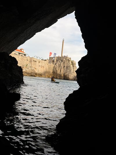 The image depicts a view from within a dark cave, looking out towards a sunlit scene of a stone fortress and the sea. The foreground is dominated by the rough, dark textures of the cave walls, which frame the opening.
Beyond the cave, a weathered stone fortress with crenellated walls and arched windows stands against a bright, overcast sky. Two flags, one red and one blue with stars, are visible flying from the fortress. A tall wooden pole extends from the top of the fortress, with what appears to be a decorative element at its apex.
In the water, a person on a paddleboard is visible near the fortress walls. The sea is relatively calm, with sunlight reflecting off its surface, creating a shimmering pattern. The water appears to be a deep blue. The overall scene suggests a historic coastal location, possibly a fortified city. The lighting indicates it is daytime, with the sun obscured by clouds. The presence of the fortress and the sea suggest a location with historical significance and natural beauty. No visible text is present in the image.