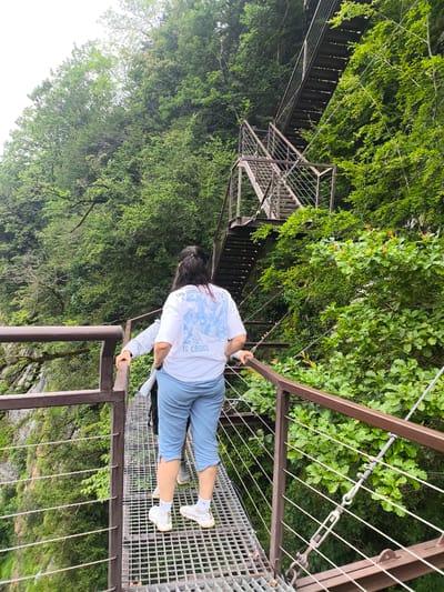 Two individuals are walking on a metal walkway suspended over a deep gorge covered in lush green foliage. The walkway has a dark brown metal railing and a grated metal surface. Behind them, a steep metal staircase ascends up the side of the gorge, also surrounded by dense trees. The person in the foreground is wearing a white t-shirt with a blue graphic and light blue capri pants. The second person, partially visible, is wearing a light-colored top and dark pants. The environment suggests a natural, mountainous area. This location is likely the Okatse Canyon, near the village of Satsiskvilo, Georgia, known for its suspension bridge and walkways.