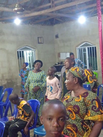 This indoor photograph captures a diverse group of people, including adults and children, gathered in a simple community space or perhaps a church in Nando-Ikem, Nigeria. The scene is brightly lit by artificial light bulbs and a ceiling fan is visible overhead, suggesting the event is taking place in the evening or at night.
In the foreground, the face of a young boy is partially visible, looking directly towards the viewer. To his right, an adult woman wearing a vibrant green, brown, and yellow patterned dress and a matching headwrap is visible, looking to her left. To the left foreground, another young boy in a black shirt with yellow patterns and black trousers (which have indistinct white text, possibly "AMIRI") is seated on a blue plastic chair, appearing to be resting or observing.
In the midground, several individuals are standing. A woman in a blue, yellow, and red patterned dress has her mouth open and is looking upwards, suggesting she is singing or speaking enthusiastically. Beside her, another woman in a similar patterned dress and a young boy in a light-colored long-sleeve shirt are also looking upwards, actively engaged in the activity.
The setting features light-colored, possibly plastered, walls. Two windows with white protective grates are visible on the back wall, along with a small, framed picture hanging between them. The ceiling structure reveals exposed wooden beams. Scattered blue plastic chairs, some occupied and some empty, indicate a communal gathering. Many adults are dressed in colorful traditional African print fabrics, adding to the cultural context of the scene in Nando-Ikem. The overall atmosphere appears to be one of communal participation and engagement.
