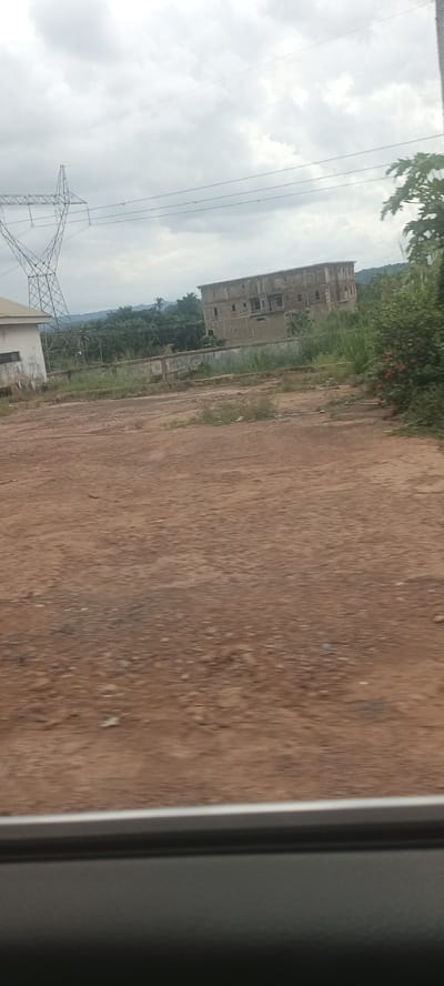 The image depicts a view in Ozalla, Nigeria, characterized by a barren, reddish-brown foreground of loose dirt and gravel. The lower part of the frame is obscured by the interior of a vehicle, suggesting the photo was taken from within. In the middle ground, there is a clearing with some sparse vegetation and a partial concrete barrier or fence. Beyond this clearing are several structures, including a partly visible white building on the left and a larger, unfinished building with multiple stories, seemingly constructed of concrete blocks, in the center. An electrical transmission tower is visible on the left side of the image, with power lines stretching across the sky. The background features a rolling landscape of green vegetation and hills under an overcast sky. No people or interactions are visible in the frame.