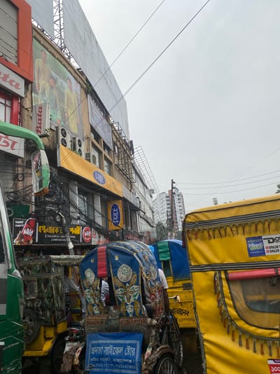 This image captures a street scene in Narayanganj, Bangladesh. The foreground features a cluster of vehicles, including auto-rickshaws and rickshaws, identifiable by their yellow and blue canopies. The vehicles are adorned with decorative elements and textual information in Bengali.

Behind the vehicles rises a building adorned with multiple advertisements and signage, including brands like "Pride" and "Bata", along with local businesses and advertisements in Bengali. The building's facade is cluttered with air conditioning units and a dense network of tangled electrical wires.

The sky above is overcast, suggesting a cloudy day. The image, though angled, provides a cluttered, realistic view of an urban scene in Narayanganj.