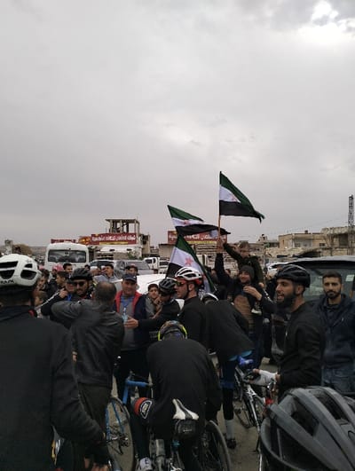 A group of cyclists and onlookers gather in a street, some holding Syrian flags. The cyclists are wearing helmets and cycling gear, while the other people are dressed in casual clothing. A man holds a child on his shoulders. The scene appears to be a celebration or rally, with people interacting and possibly shaking hands. Vehicles are parked nearby, and buildings are visible in the background under an overcast sky.